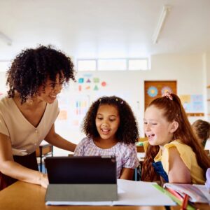 Happy young female teacher explaining to schoolgirls using digital tablet, laughing