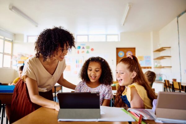 Happy young female teacher explaining to schoolgirls using digital tablet, laughing